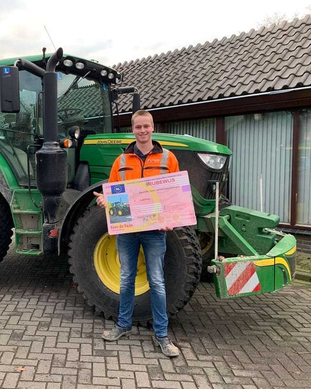 Geslaagde cursist in oranje werkkleding met rijbewijsbord voor de bumper van een groene lestrekker