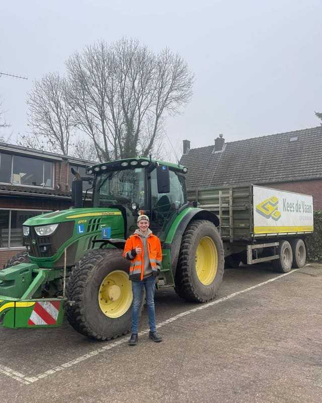 Geslaagd voor het trekkerrijbewijs: cursist in oranje werkjas steekt duim op voor de lestrekker met aanhangwagen