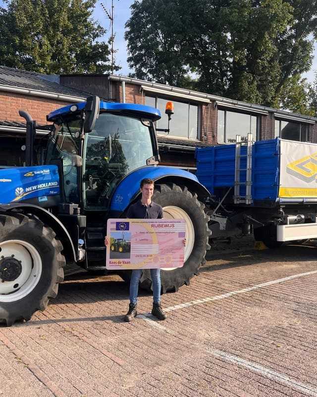Geslaagd voor het trekkerrijbewijs: cursist met rijbewijsbord voor een blauwe lestrekker met blauwe kipper