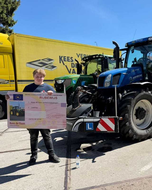 Geslaagd voor het trekkerrijbewijs: cursist met rijbewijsbord voor een gele vrachtwagen van Kees de Vaan en een blauwe lestrekker