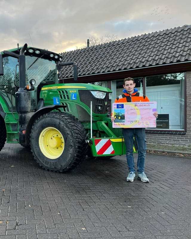 Geslaagd voor het trekkerrijbewijs: cursist in oranje jas met rijbewijsbord naast een groene lestrekker