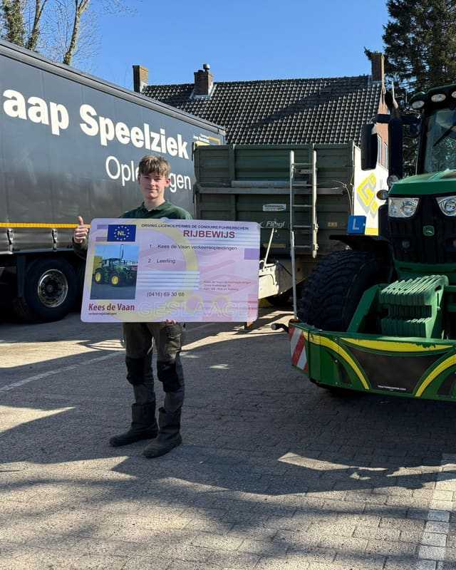 Geslaagde cursist met rijbewijsbord tussen een vrachtwagen en een groene lestrekker