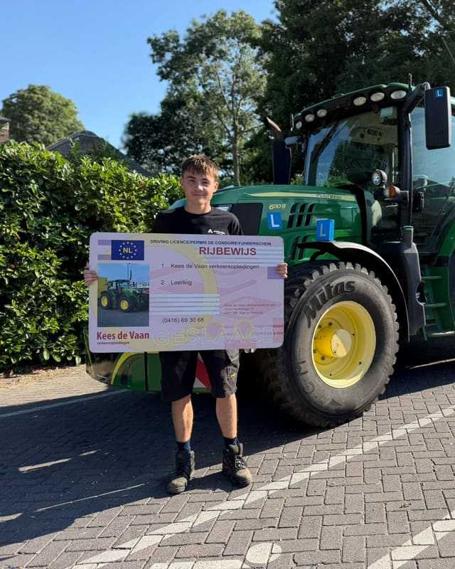 Geslaagde cursist in korte broek met groot rijbewijsbord bij een groene lestrekker